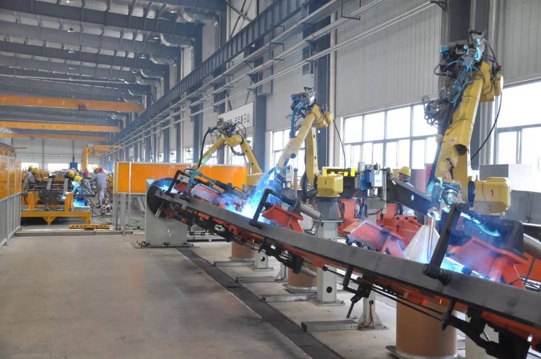 Robotic welding line in a spacious factory with yellow arms welding metal on a tilted conveyor; workers are seen in the background.