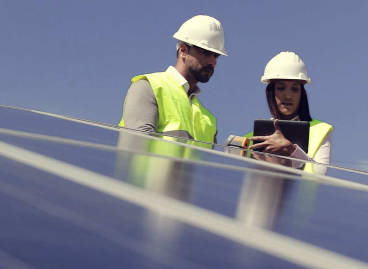 Two construction workers in hard hats and high-visibility vests inspect a row of solar panels, one holding a tablet and the other monitoring with a device.